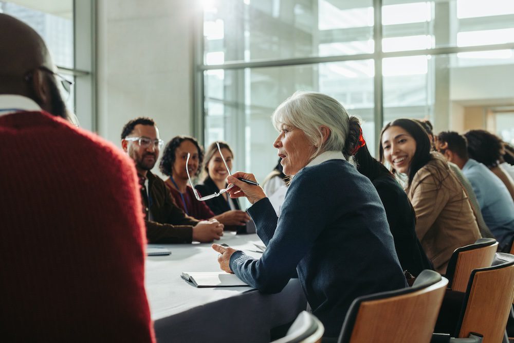 Diverse group of business professionals in a meeting with a woman speaking. Colleagues engage in a collaborative discussion in a modern conference room.