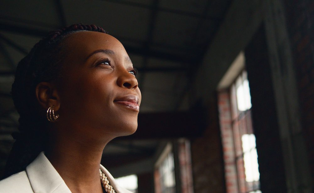 Portrait of a confident Black woman in a modern industrial office setting, gazing thoughtfully. The image captures feelings of ambition and introspection with brick walls and large windows in the background.